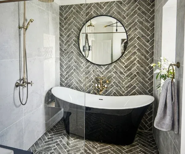 Herringbone tile accent wall behind a black freestanding tub and a round mirror