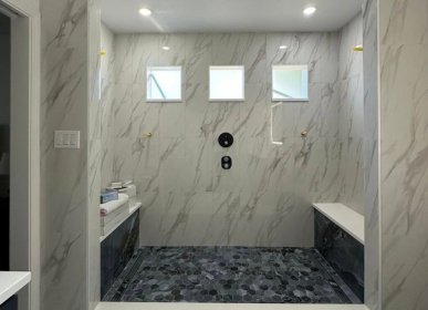Spacious walk-in shower remodel in Wilmington, NC featuring marble-look walls, mosaic tile flooring, built-in benches, and modern black fixtures.