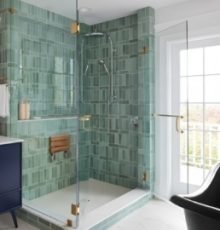 Modern bathroom remodel in Wilmington, NC featuring a glass-enclosed shower with green tile, brass hardware, and a fold-down shower seat.